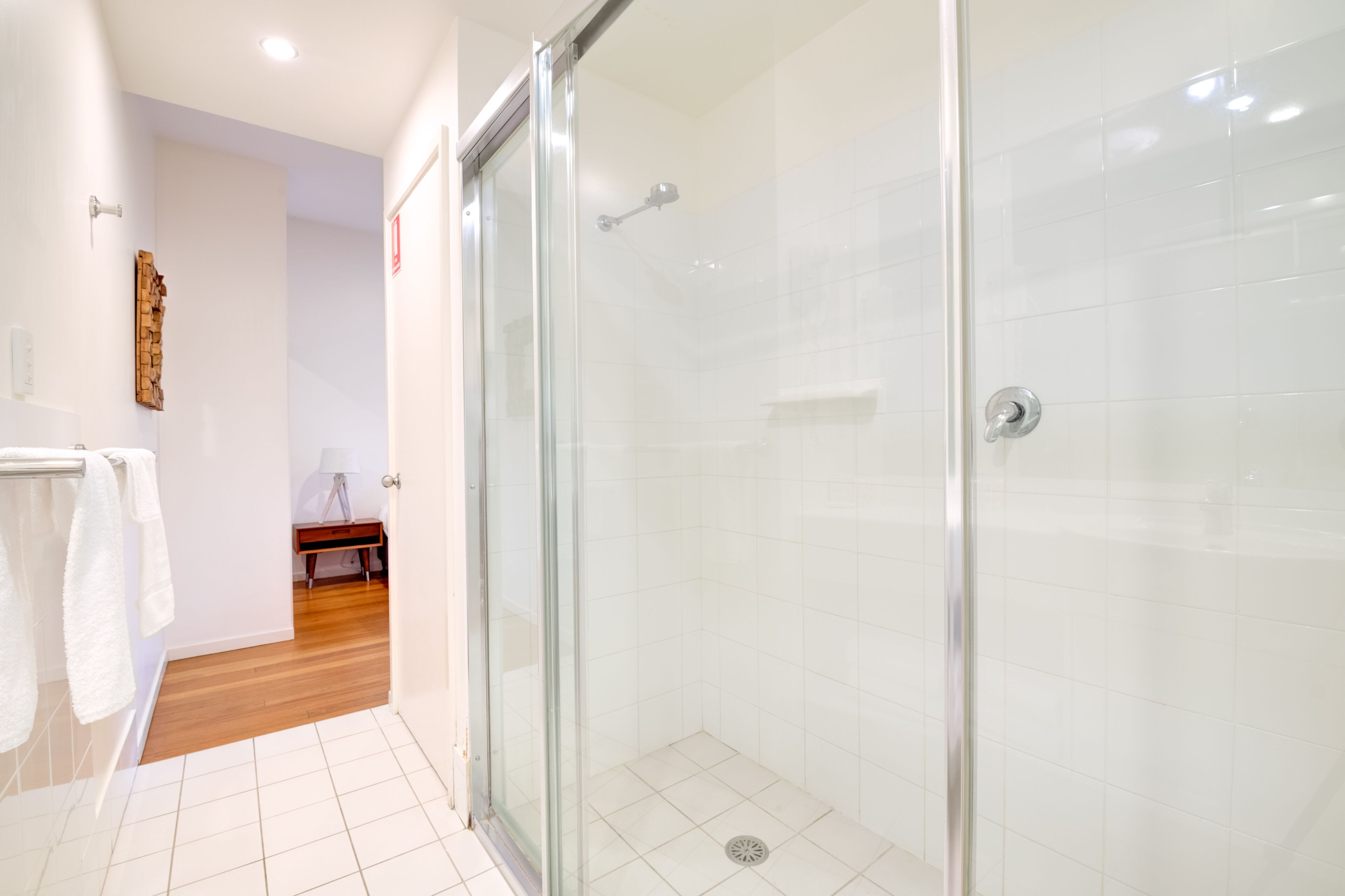 A modern bathroom with a glass shower enclosure, white tiles, and a wooden floor.