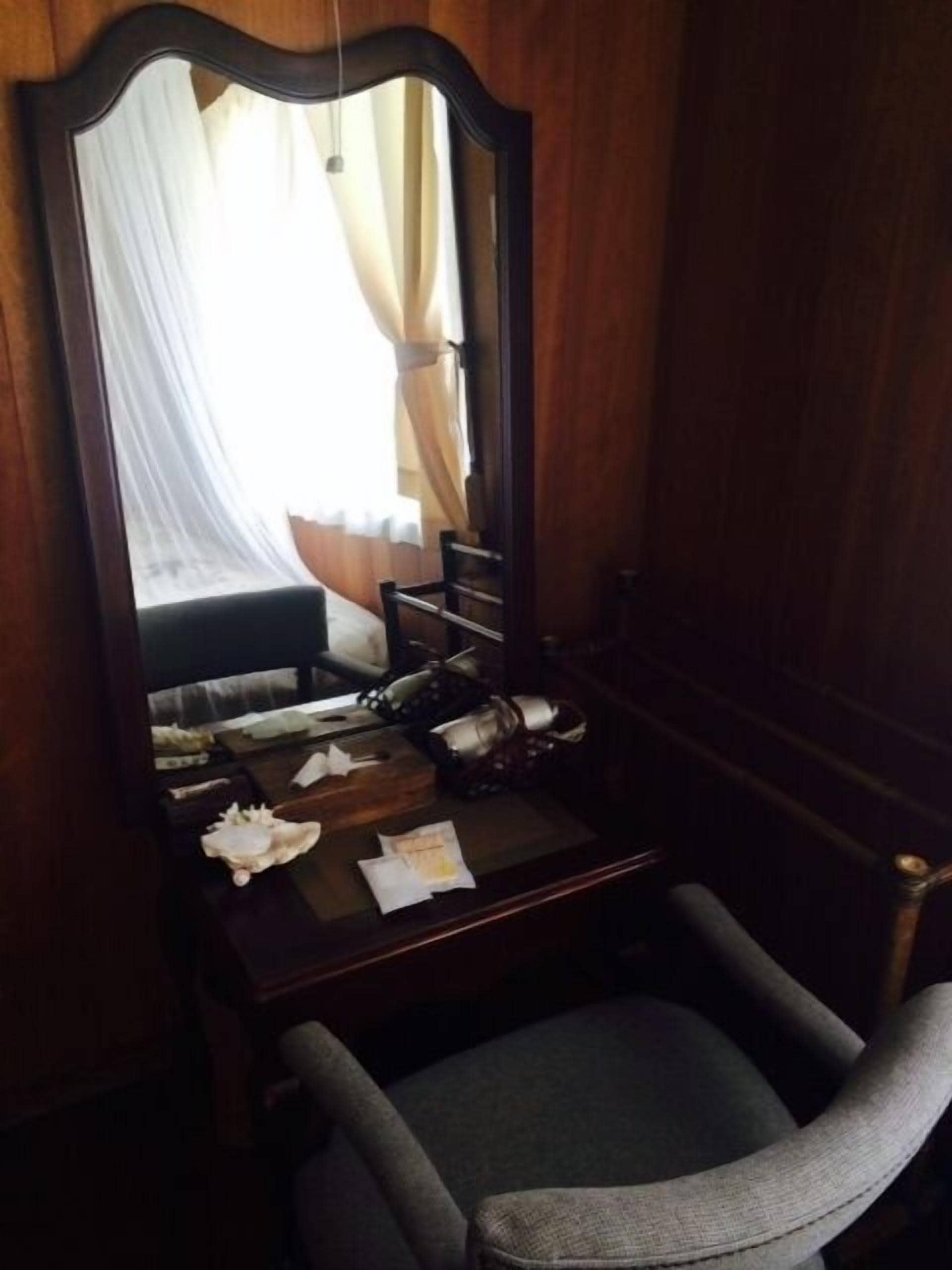 A wooden-framed mirror with a bed and curtains in the background, a desk with a chair, and a small table with personal items.