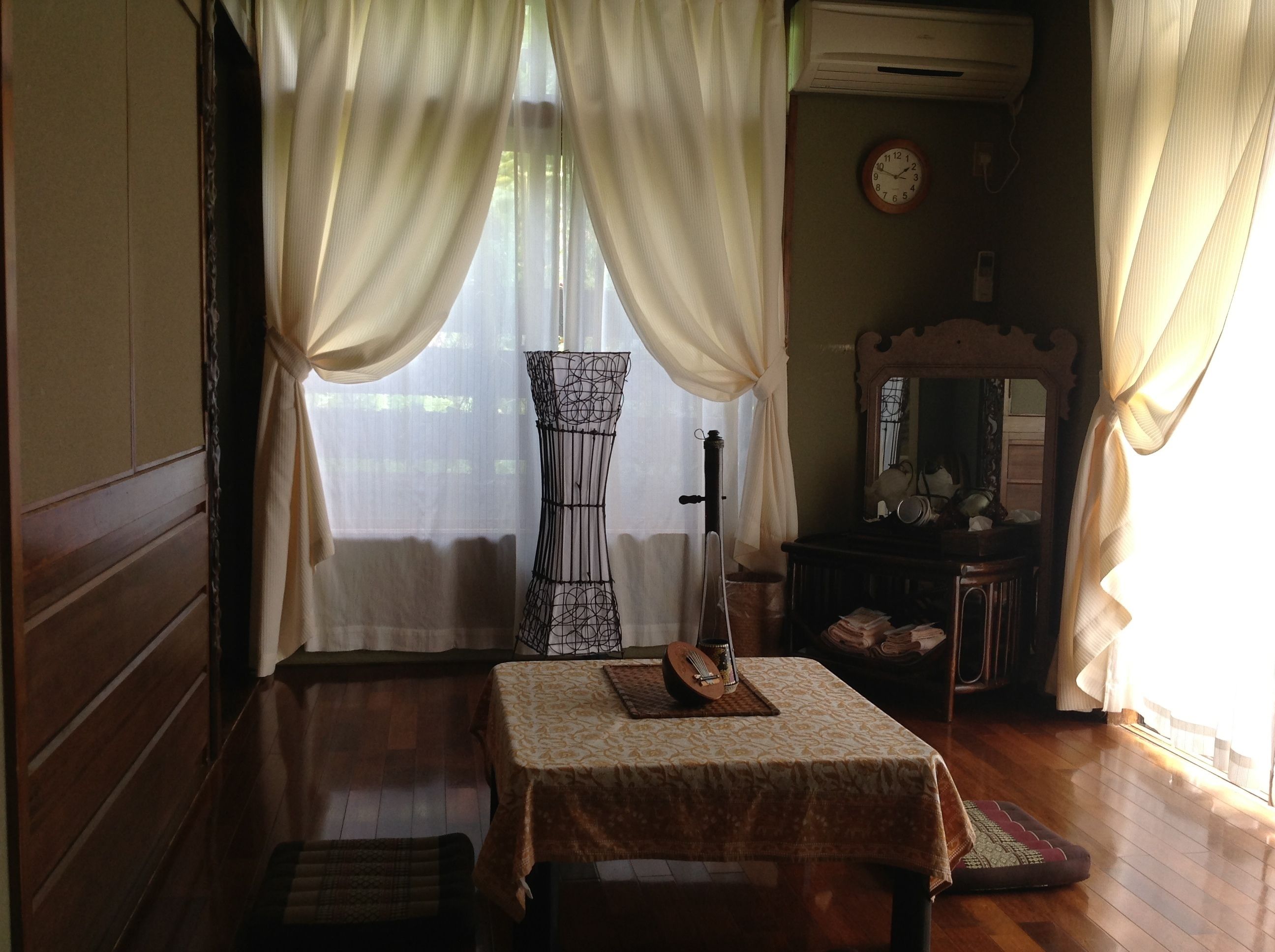 A room with a wooden floor, a table with a patterned tablecloth, a chair, a small stool, a wall clock, a mirror, and a window with sheer curtains.