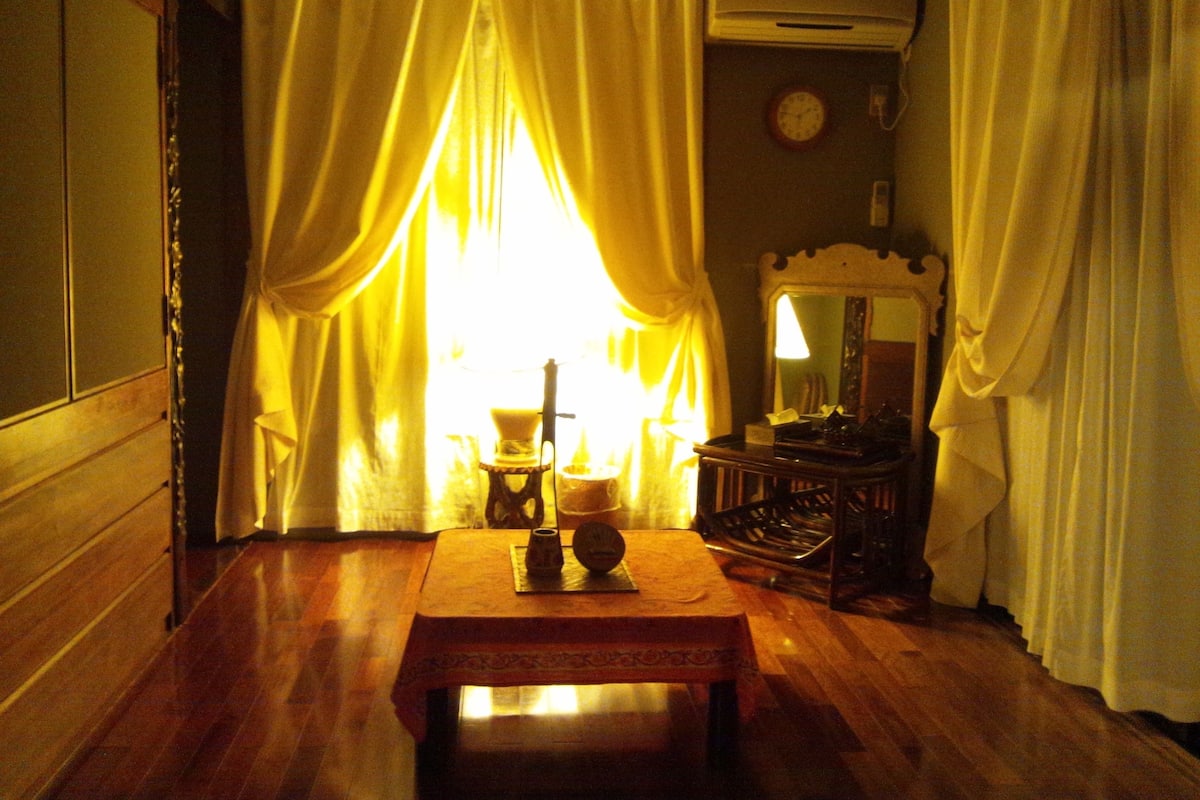 Una habitaci ón con piso de madera, una mesa con lámpara, un reloj en la pared y cortinas corridas a un lado.