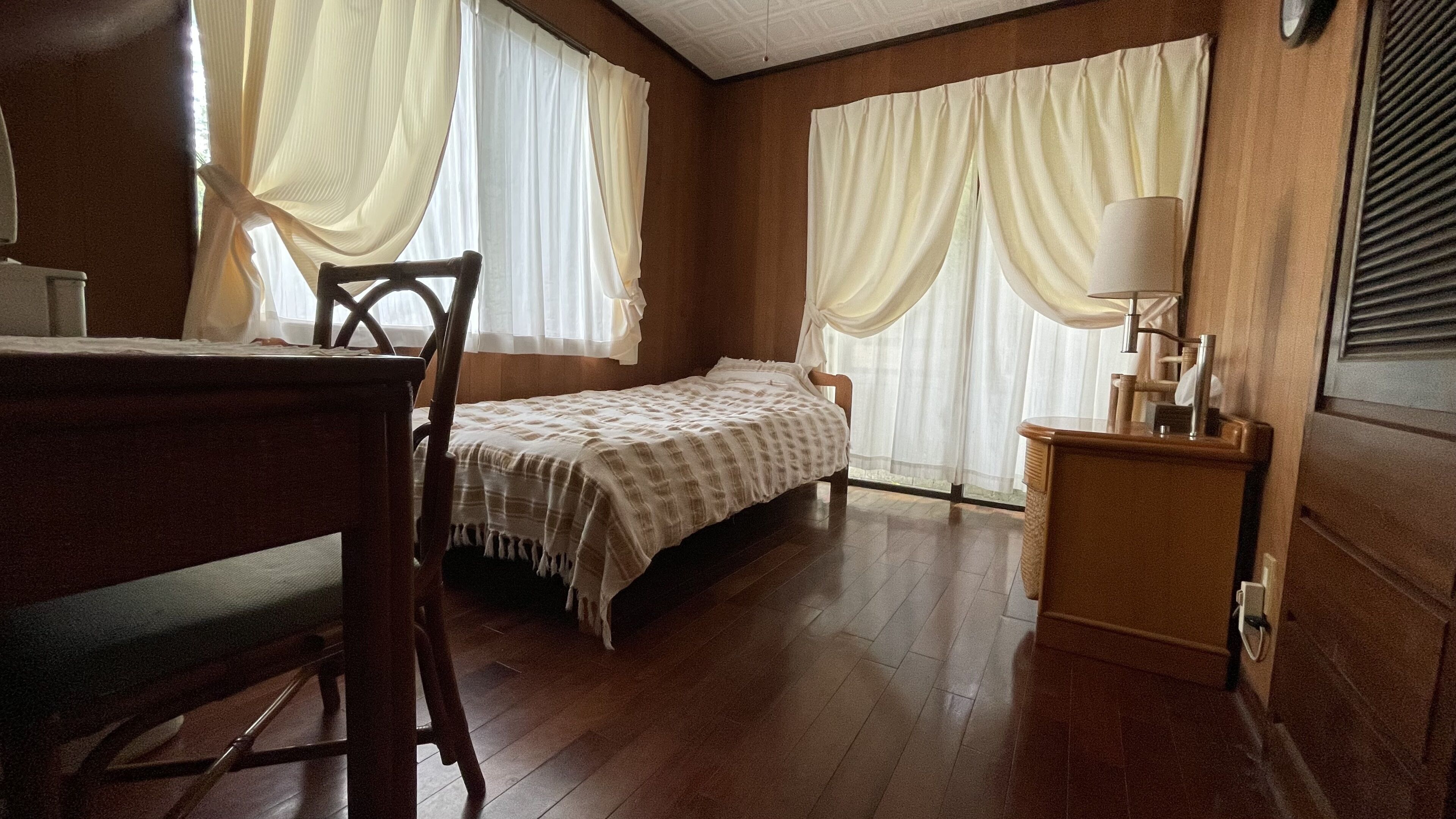 A room with a wooden floor, a bed with a striped bedspread, a chair, a bedside table with a lamp, and curtains on the windows.