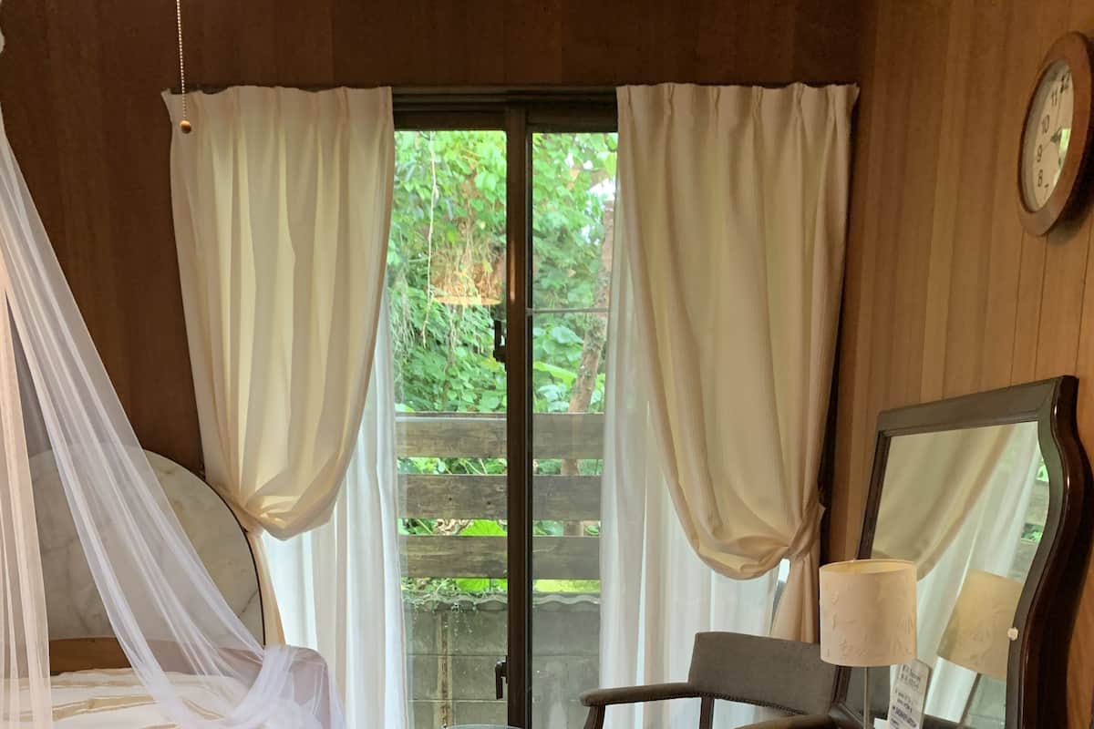 Una habitación con cama, una silla, una mesa con lámpara, un espejo y una ventana con cortinas.