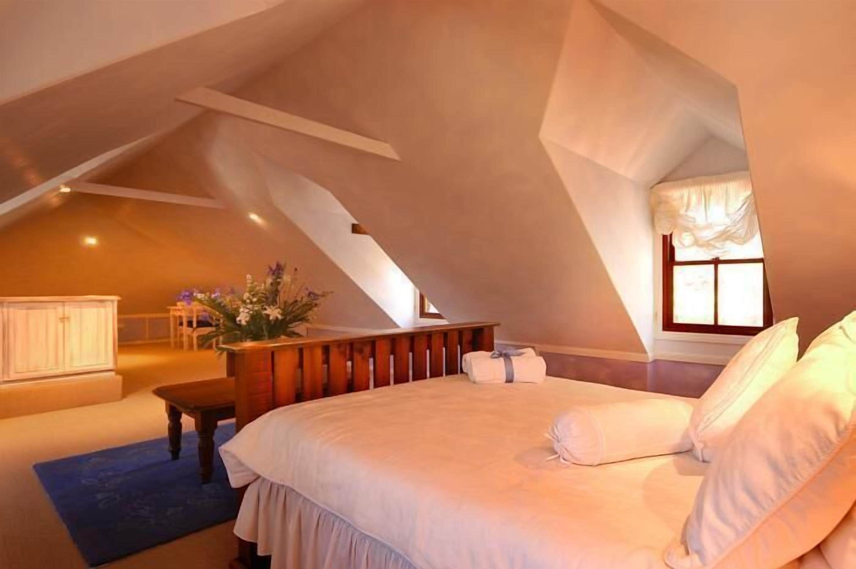 A bedroom with a large bed, a wooden bench, a small table, and a window with curtains.