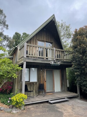 A wooden cabin with a balcony, a covered porch, and a small outdoor seating area. - Daylesford Spa Accommodation (Daylesford)