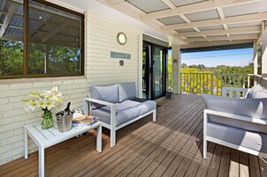 Un espacio al aire libre techado con una mesa blanca, dos sofás grises, un florero con flores y un cartel con el número del apartamento.