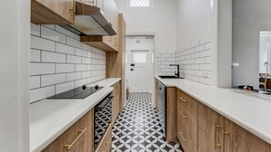 A narrow kitchen with white subway tiles, wooden cabinets, and a black and white patterned floor. - Wyatt Guest House (Mount Gambier)