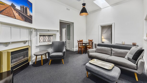A modern living room with a grey sofa, a small round table, and a chair. There is a painting on the wall and a ceiling light fixture. - Wyatt Guest House (Mount Gambier)