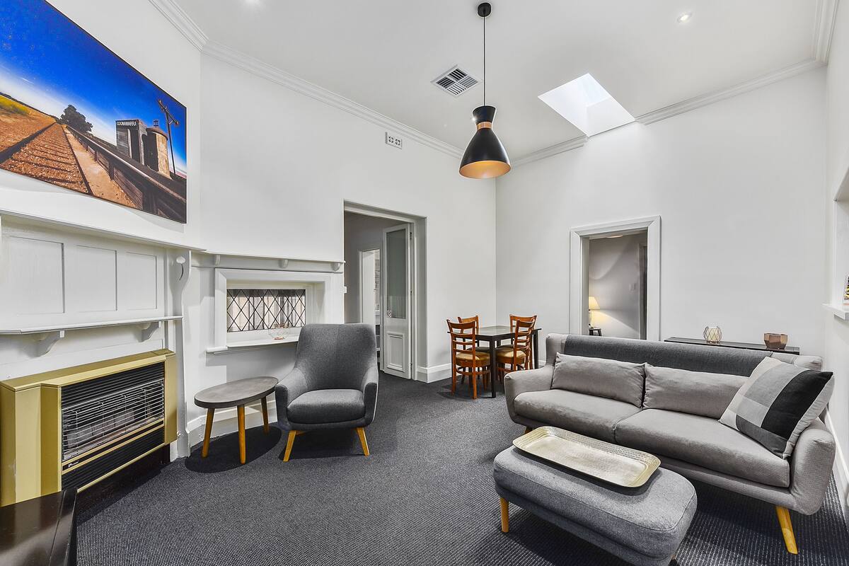 A modern living room with a grey sofa, a small round table, and a chair. There is a painting on the wall and a ceiling light fixture.
