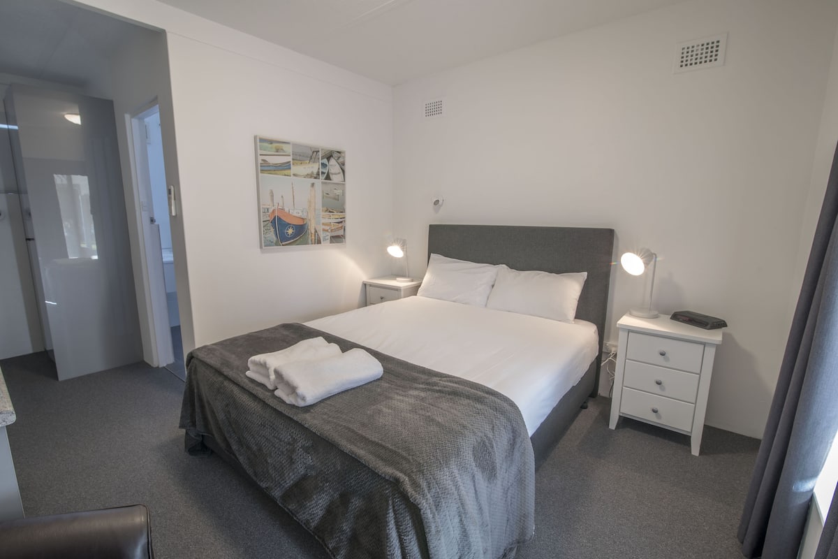 Ein ordentlich bezogenes Bett mit grauer Decke und weißen Kissen, ein Nachttisch mit einer Lampe und ein an der Wand hängendes Kunstwerk.