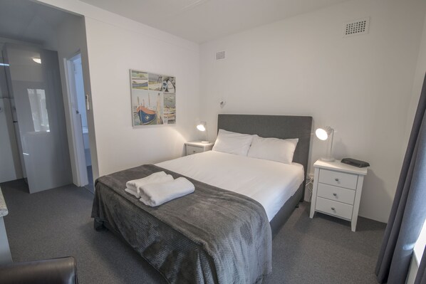 A neatly made bed with a grey blanket and white pillows, a bedside table with a lamp, and a wall-mounted artwork.