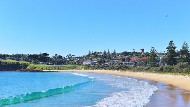 A coastal beach scene with clear blue waters, a sandy shore, and a town in the background. - Bermagui Motor Inn (Bermagui)
