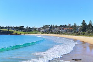 A coastal beach scene with clear blue waters, a sandy shore, and a town in the background.