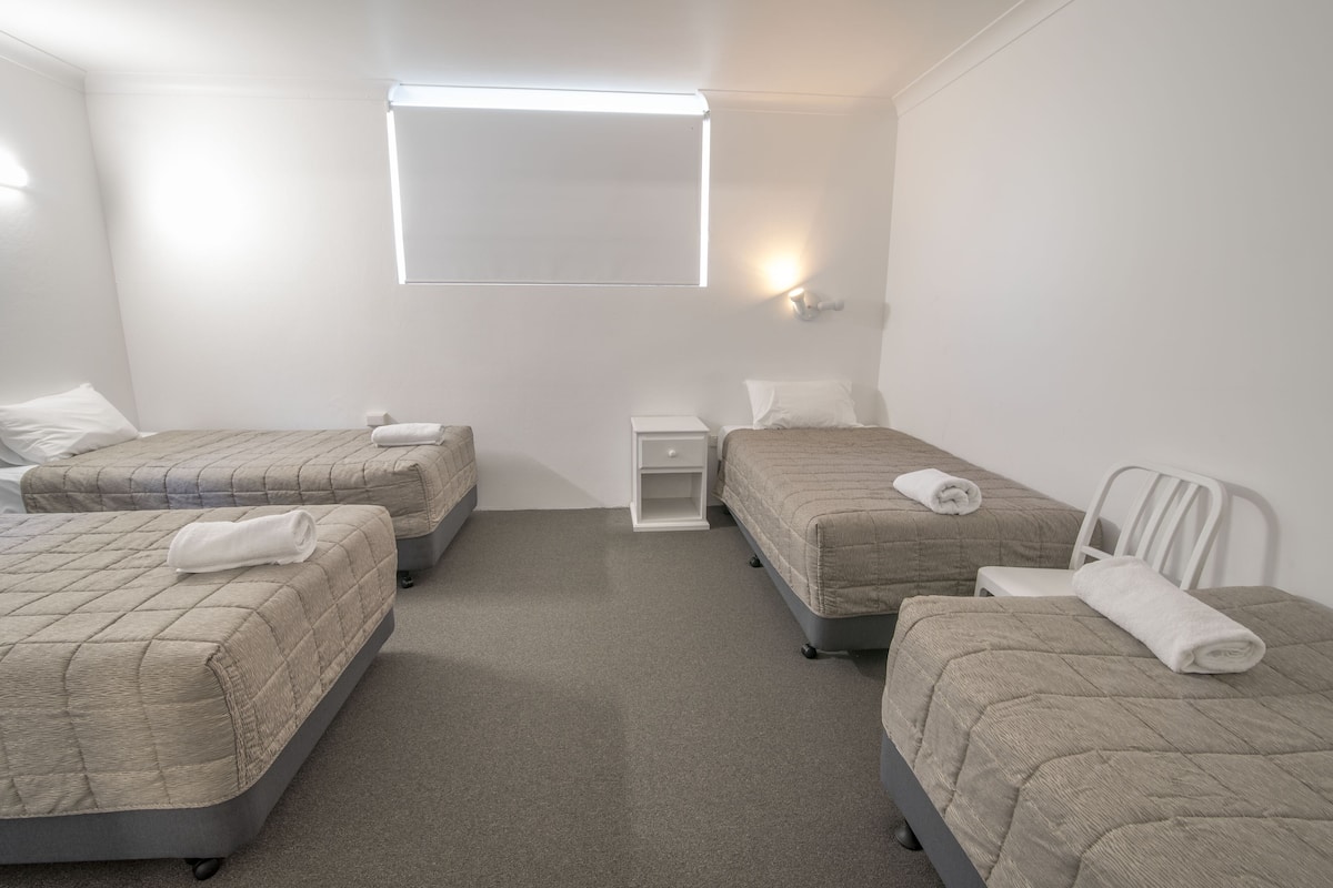 Ein Hotelzimmer mit drei Betten, einem kleinen Nachttisch, einer Wandleuchte und einem Fenster.