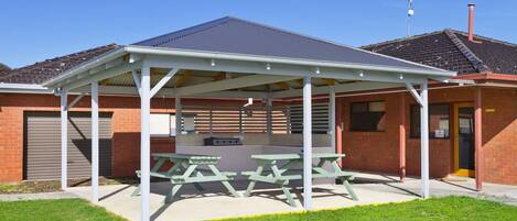 A covered picnic area with picnic tables and a barbecue grill, adjacent to a brick building with a garage.