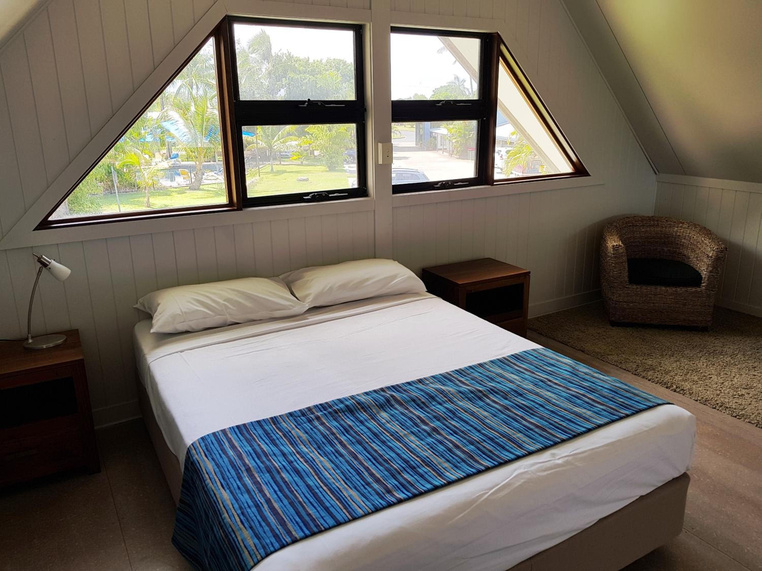 A bedroom with a bed, two bedside tables, a lamp, a chair, and a window with a view of greenery.