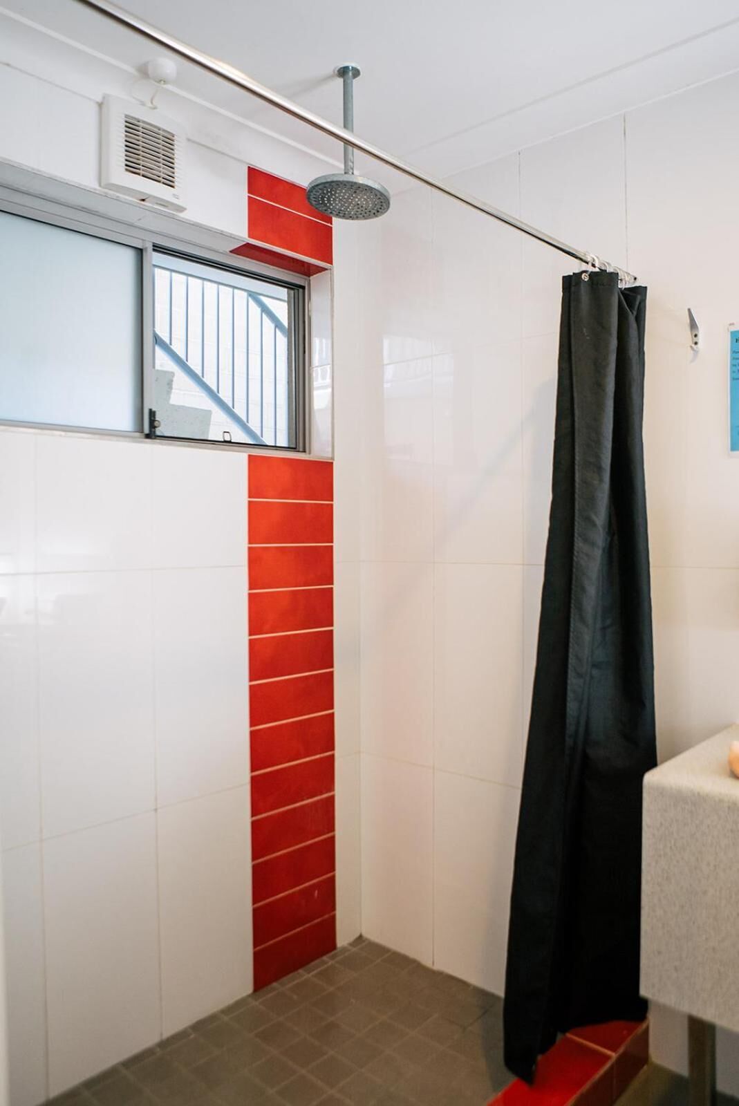 A small, modern bathroom with a shower, a window with bars, and a red-tiled accent wall.