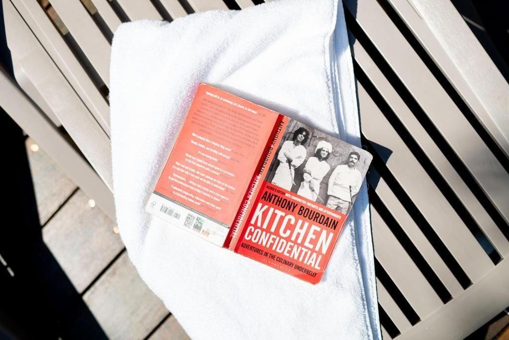 A book titled 'Kitchen Confidential' by Anthony Bourdain placed on a white towel on a bench.