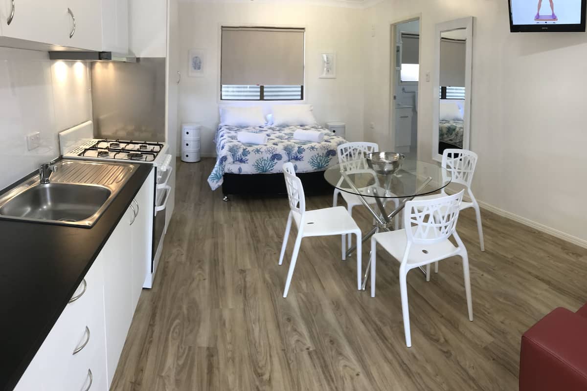 Een kleine hotelkamer met een kitchenette, een bed met een bloemenprint, een glazen eettafel met witte stoelen en een rode bank.