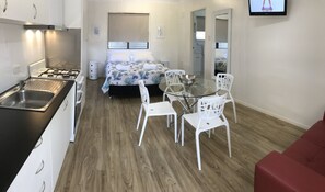 Habitación de hotel compacta con kitchenette, una cama con colcha estampada, una mesa de comedor de cristal con sillas blancas y un sofá rojo.