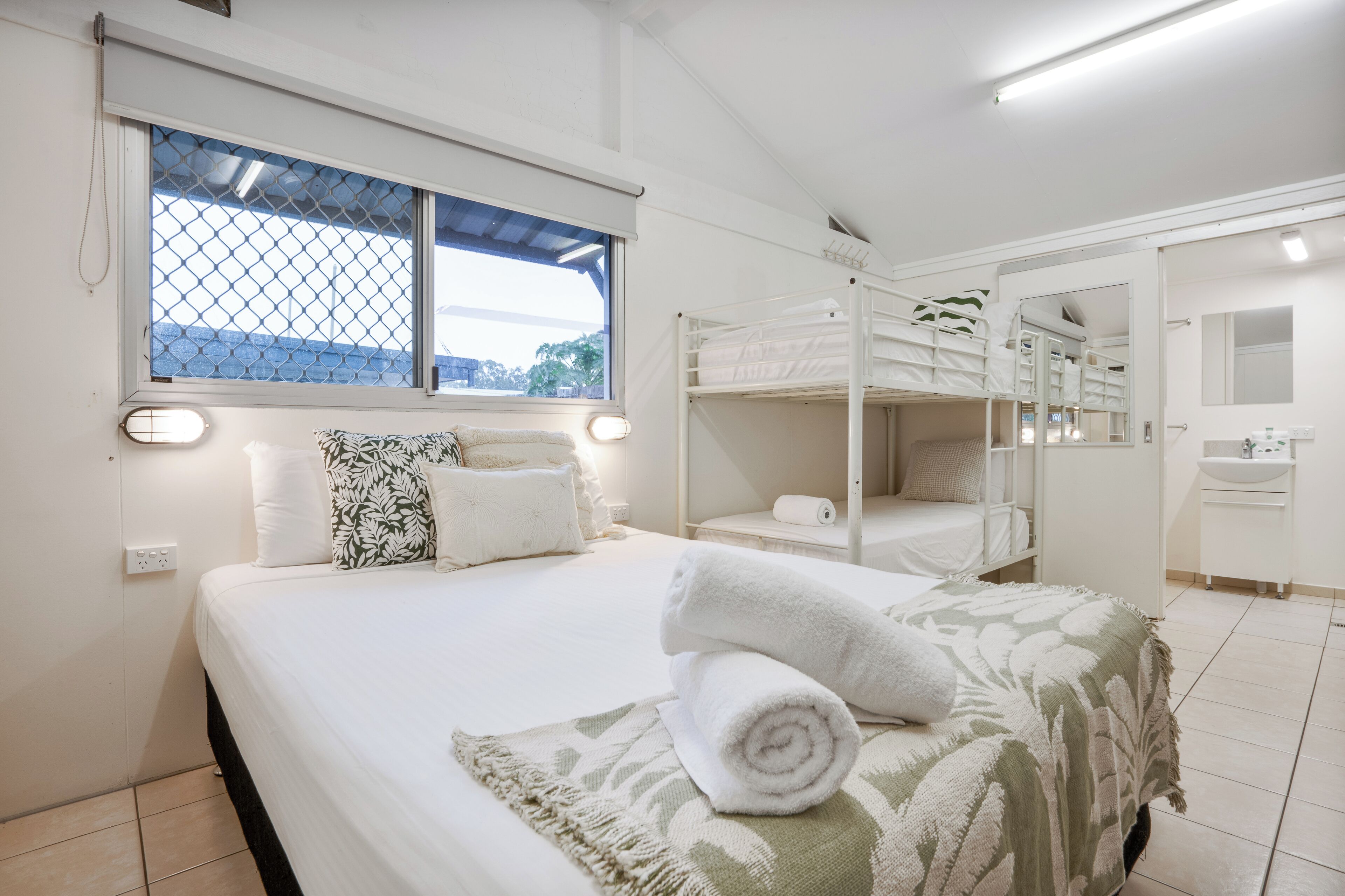 Quarto de hotel com cama, janela com vista, beliche, banheiro com pia e espelho.
