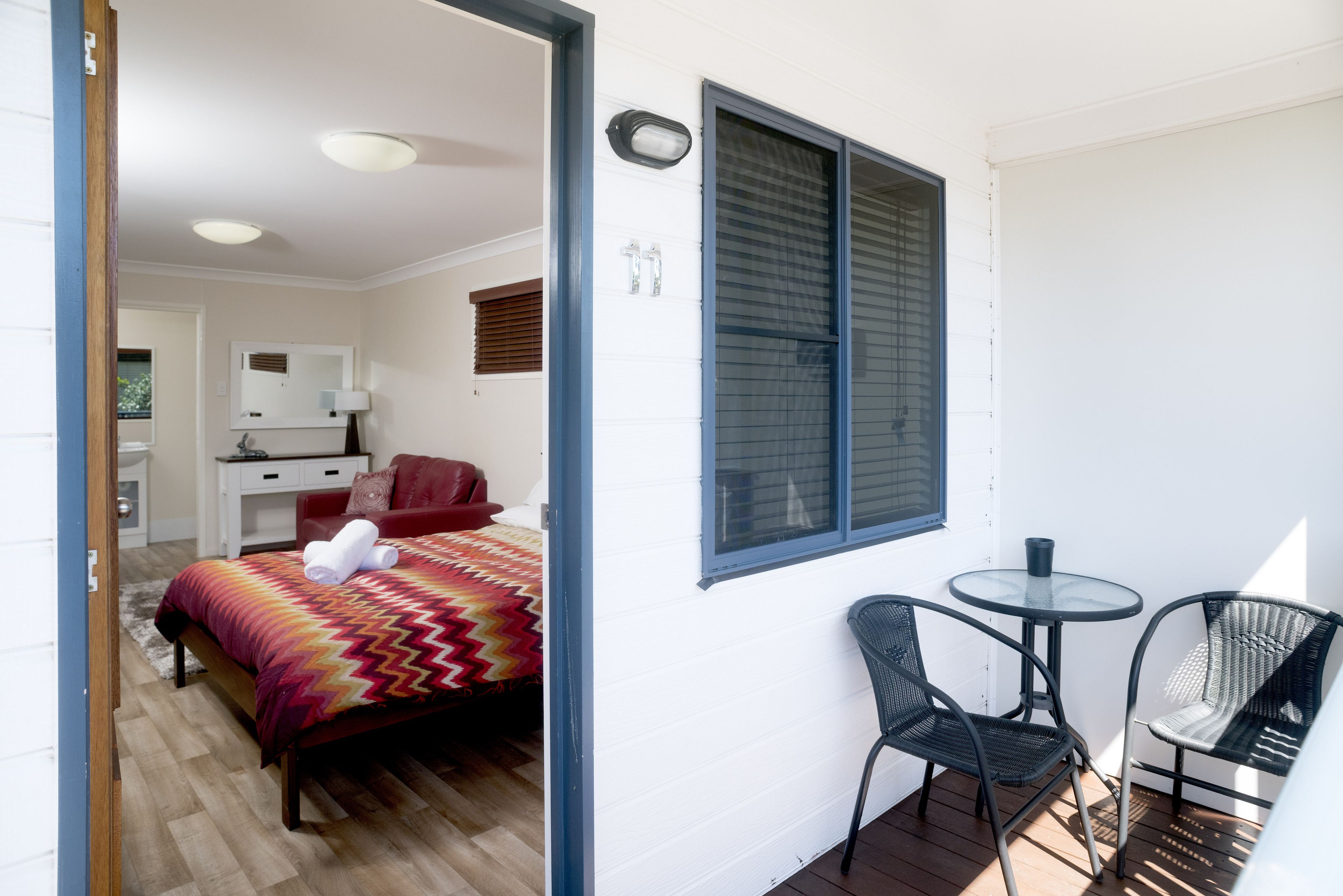 A bedroom with a bed, a desk, and a chair. There is a window with blinds and a small table with a chair.