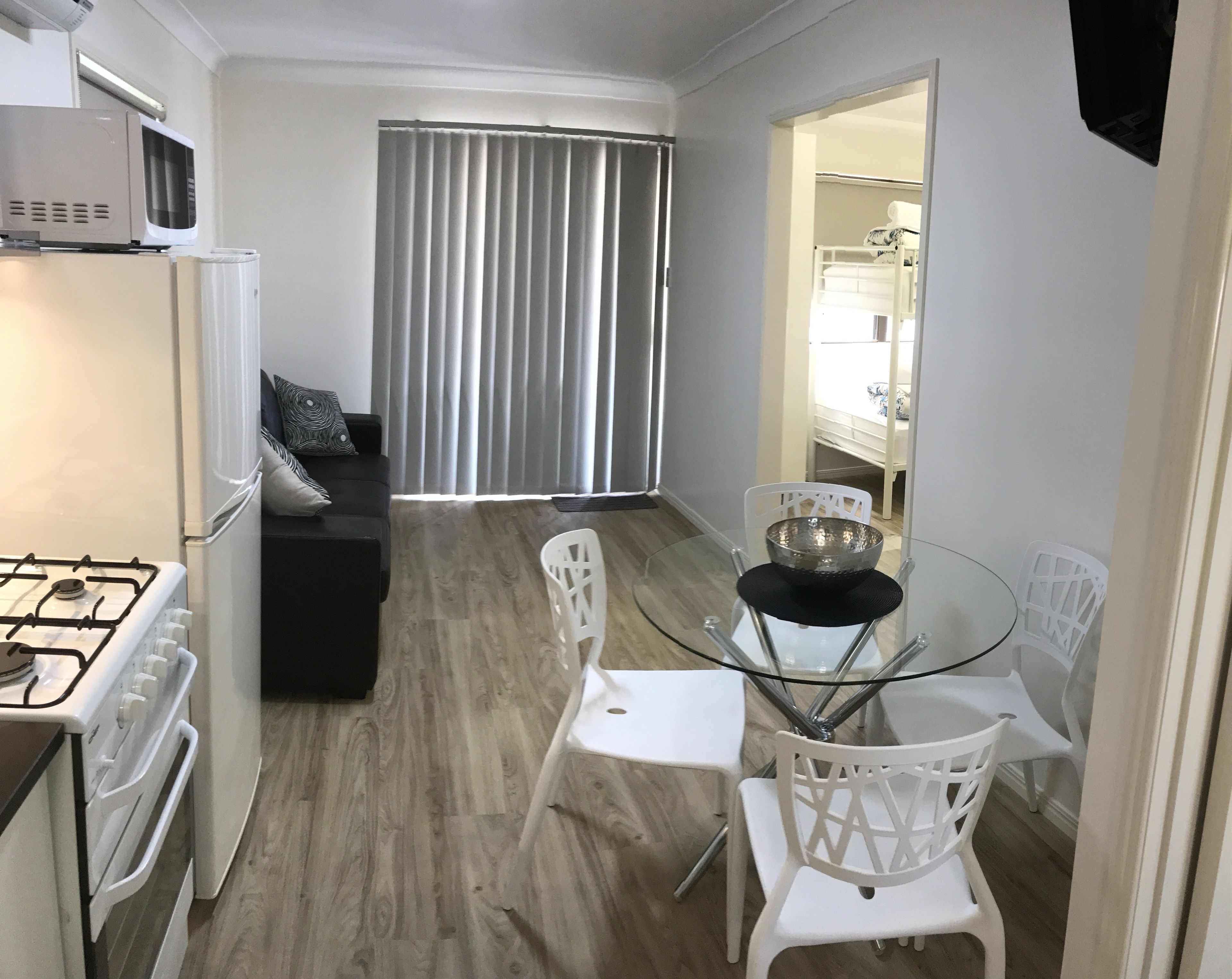 A compact kitchen with a gas stove, a microwave, and a refrigerator. There is a dining table with white chairs and a glass top. A black bowl is on the table. The room has a wooden floor and a large window with vertical blinds.