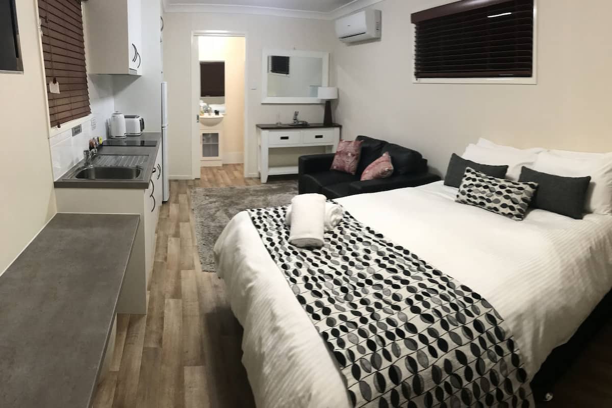 Een slaapkamer met een bed, nachtkastje en een klein houten nachttafeltje. Er is een raam met zonwering, een luchtbeheersingsunit aan de muur en een spiegel boven de dressoir.