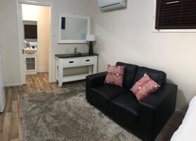 Una sala de estar con un sofá negro, dos almohadas estampadas, una consola blanca con una lámpara y un espejo encima.