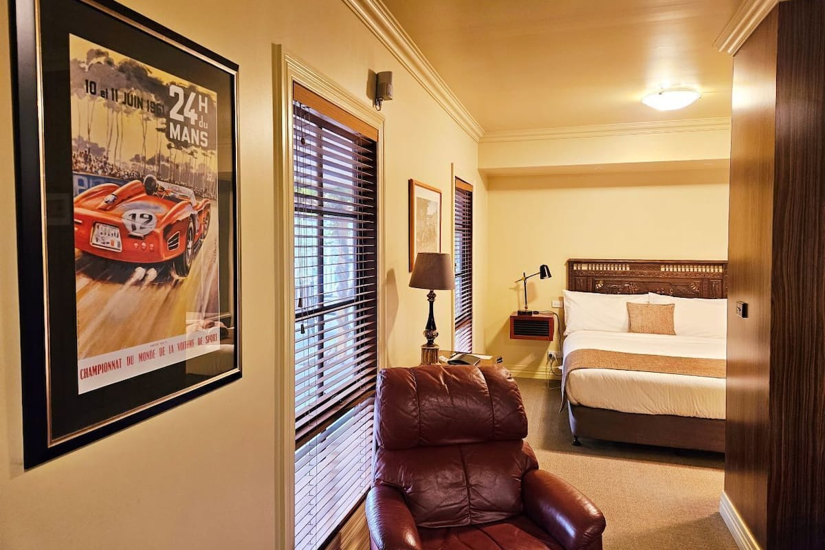 A hotel room with a bed, a leather chair, a nightstand with a lamp, and a framed poster on the wall.