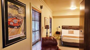 A hotel room with a bed, a leather chair, a nightstand with a lamp, and a framed poster on the wall.