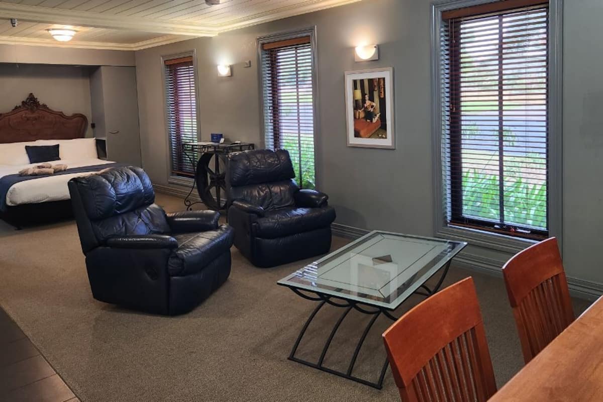 A room with a bed, two leather chairs, a glass-top table, and a wooden dining table.