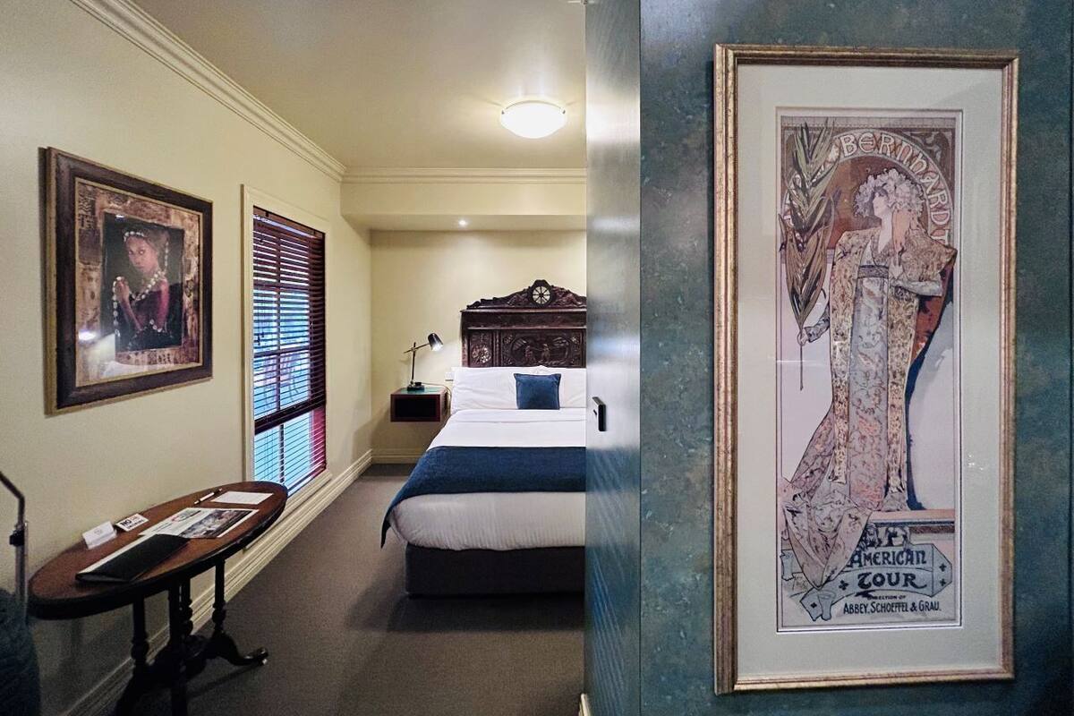 A hotel room with a bed, a nightstand, a framed picture on the wall, and a window with blinds.