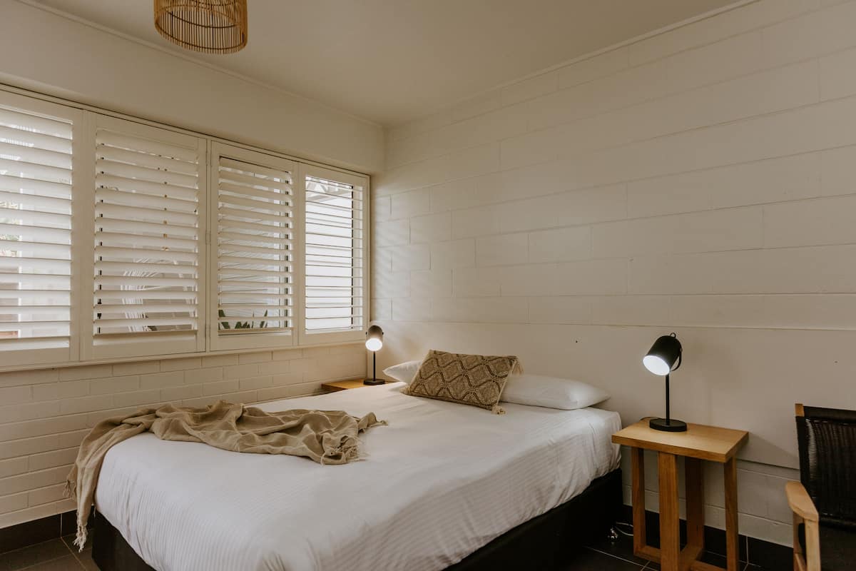 A bedroom with a bed, two bedside lamps, a window with blinds, and a chair.