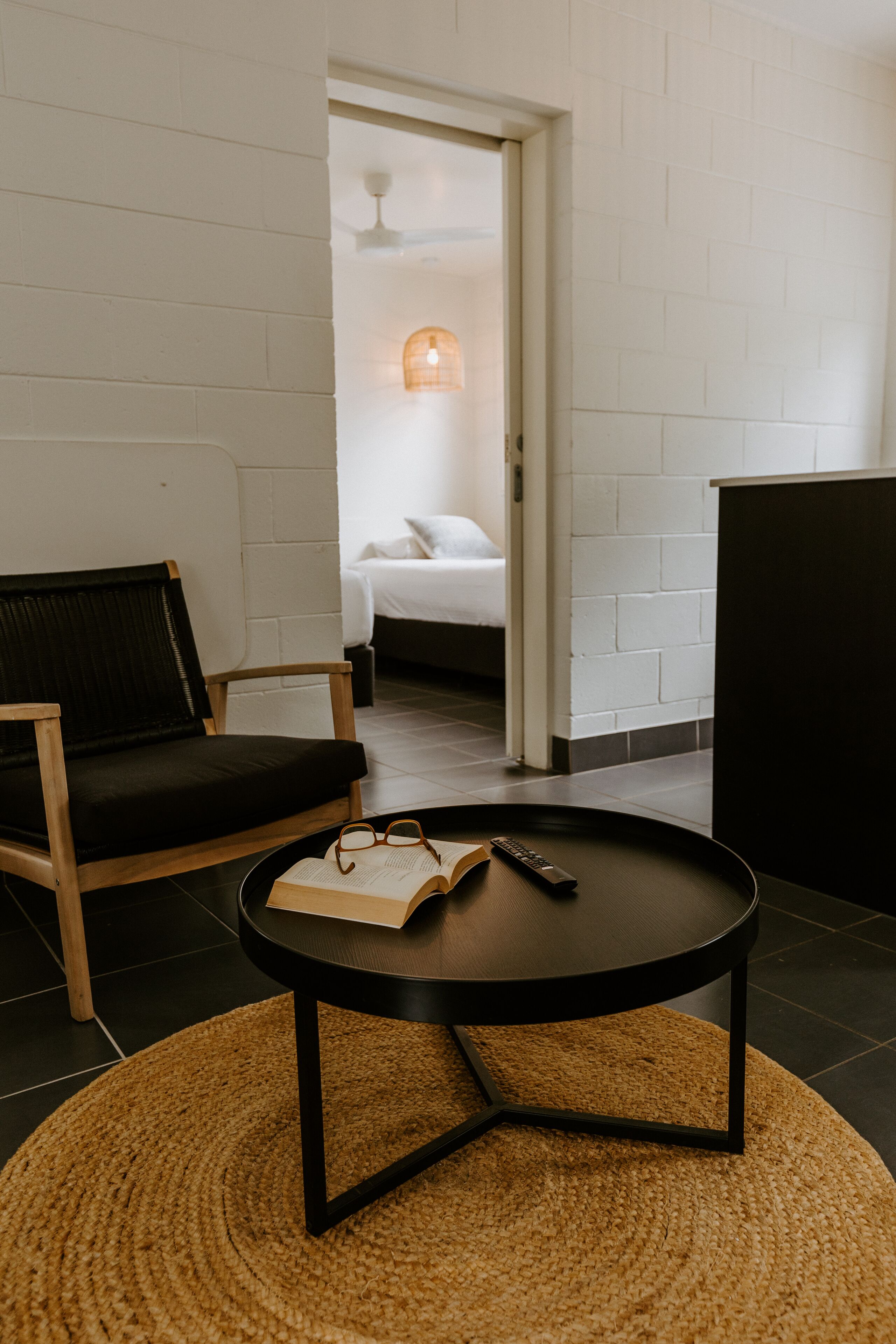 Une pièce avec un canapé noir, une table basse ronde et un tapis tissé. Au fond, on aperçoit un lit avec une literie blanche, et une lampe fixée au mur au-dessus.