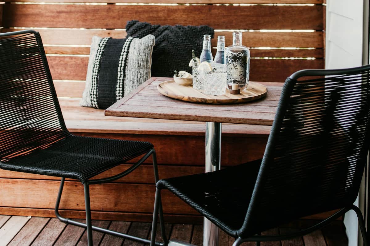 A wooden bench with a table and chairs, a white fence, and green plants in the background.