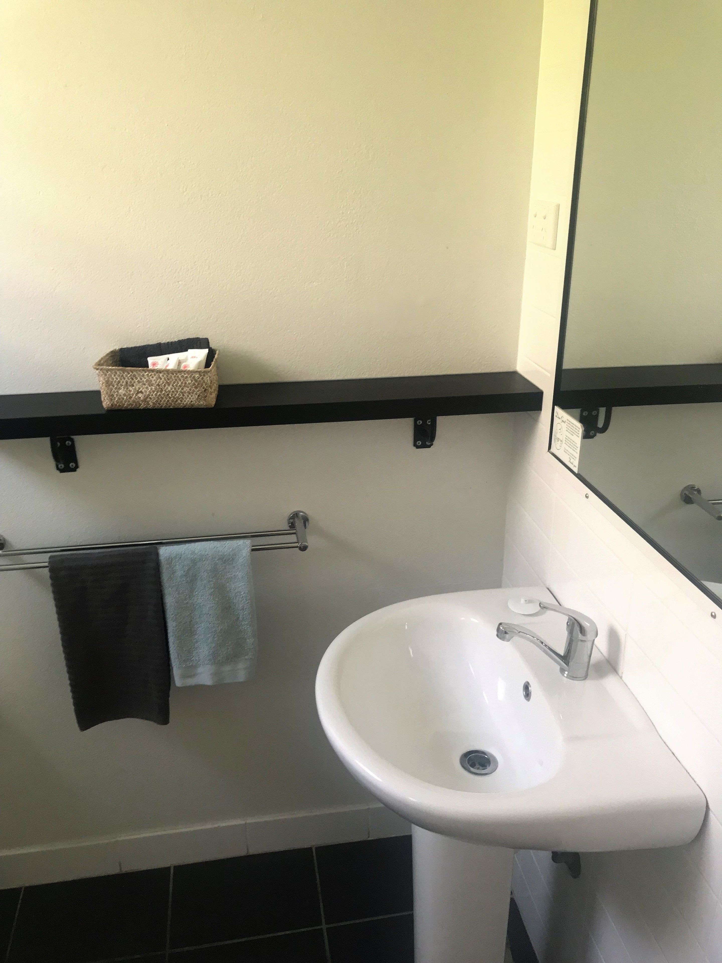 Un baño con un lavamanos blanco, un perchero con dos toallas, una cesta pequeña sobre un estante negro y un espejo sobre el lavamanos.