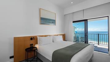 Een hotelkamer met een groot bed, een tafeltje, een stoel en een balkon met uitzicht op de oceaan.