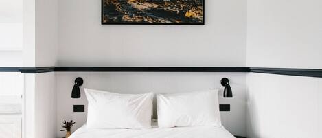 Una cama bien tendida con sábanas blancas, un cabecero negro y una fotografía enmarcada de un litoral rocoso encima.