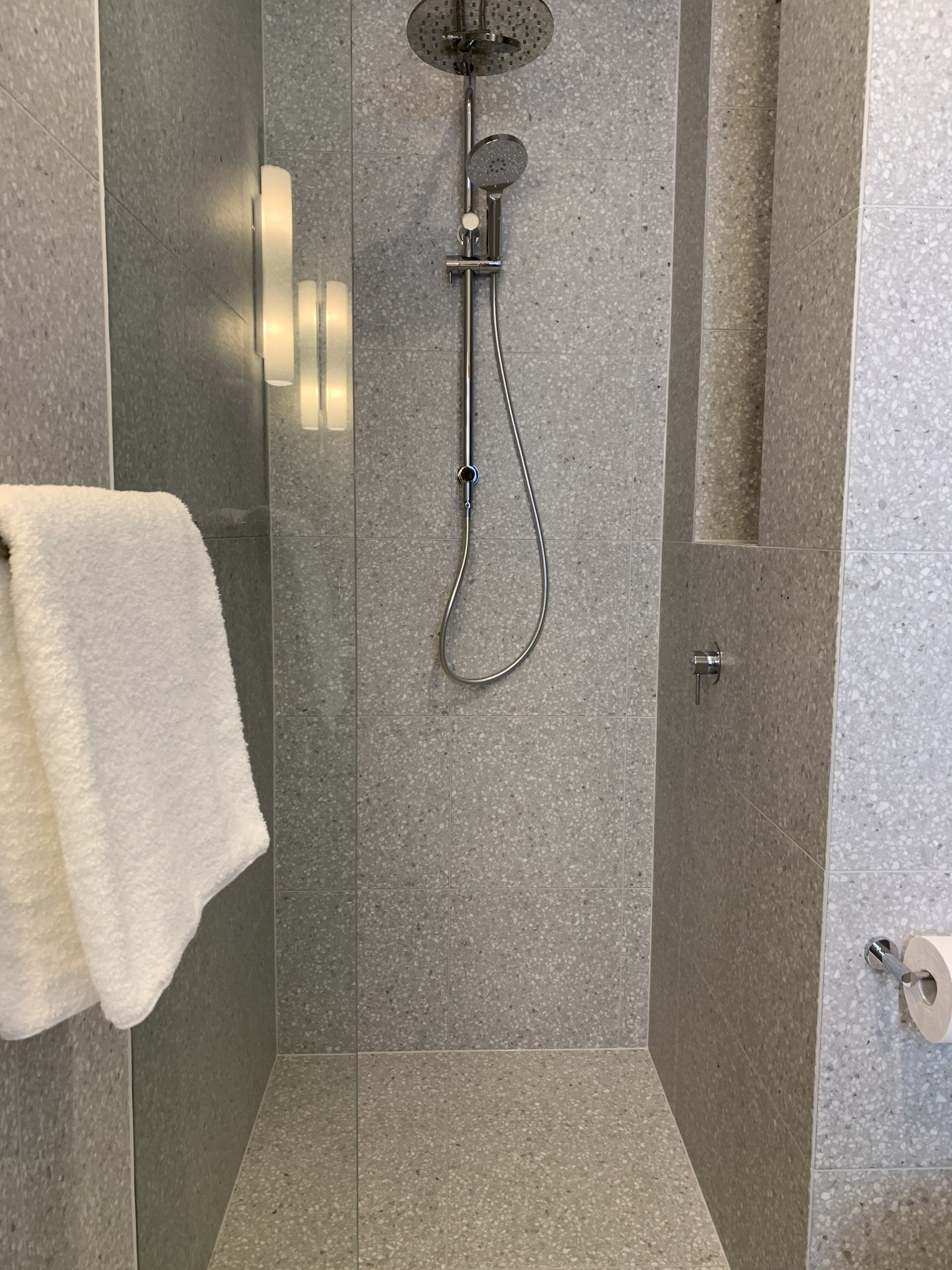 A modern shower with a handheld showerhead and a wall-mounted faucet, a white towel hanging on a rack, and a toilet paper holder on the wall.