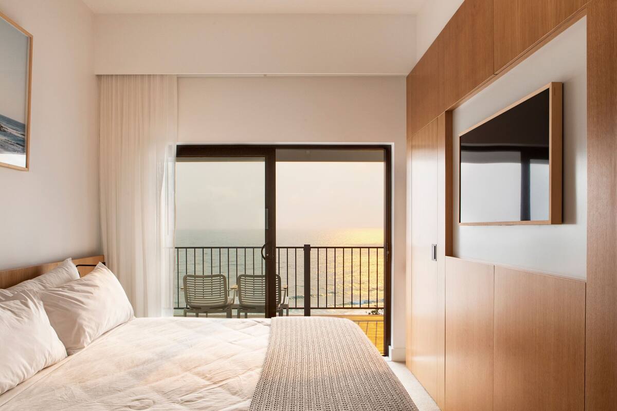 A hotel room with a large bed, a balcony with two chairs, and a view of the ocean.