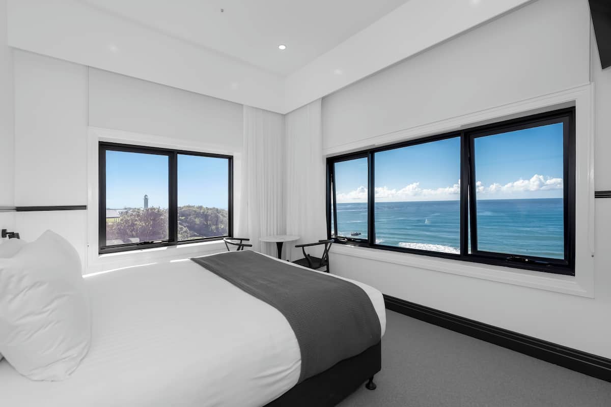 A modern hotel room with a large bed, two windows with ocean views, a small table, and a chair.