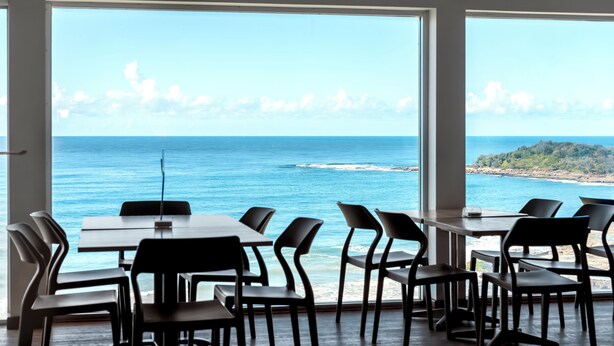 A dining area with tables and chairs facing a large window overlooking the ocean. - Pacific Hotel Yamba (Yamba)