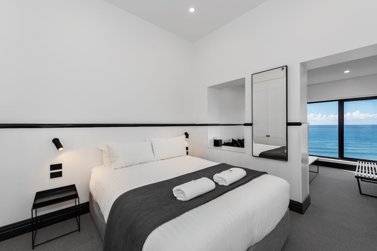 A modern hotel room with a large bed, a bedside table, a mirror, and a view of the ocean.