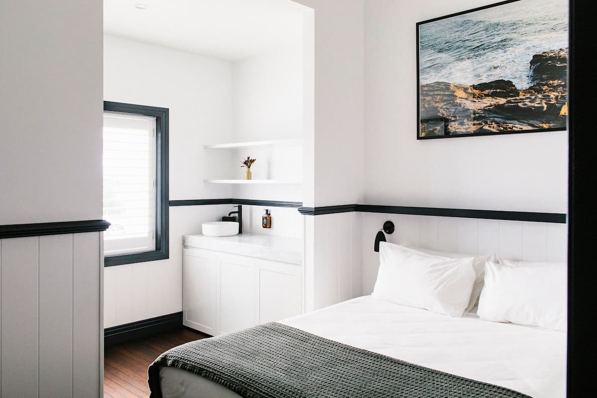 A bedroom with a bed, a window, a white dresser, and a wall-mounted shelf with a small plant.