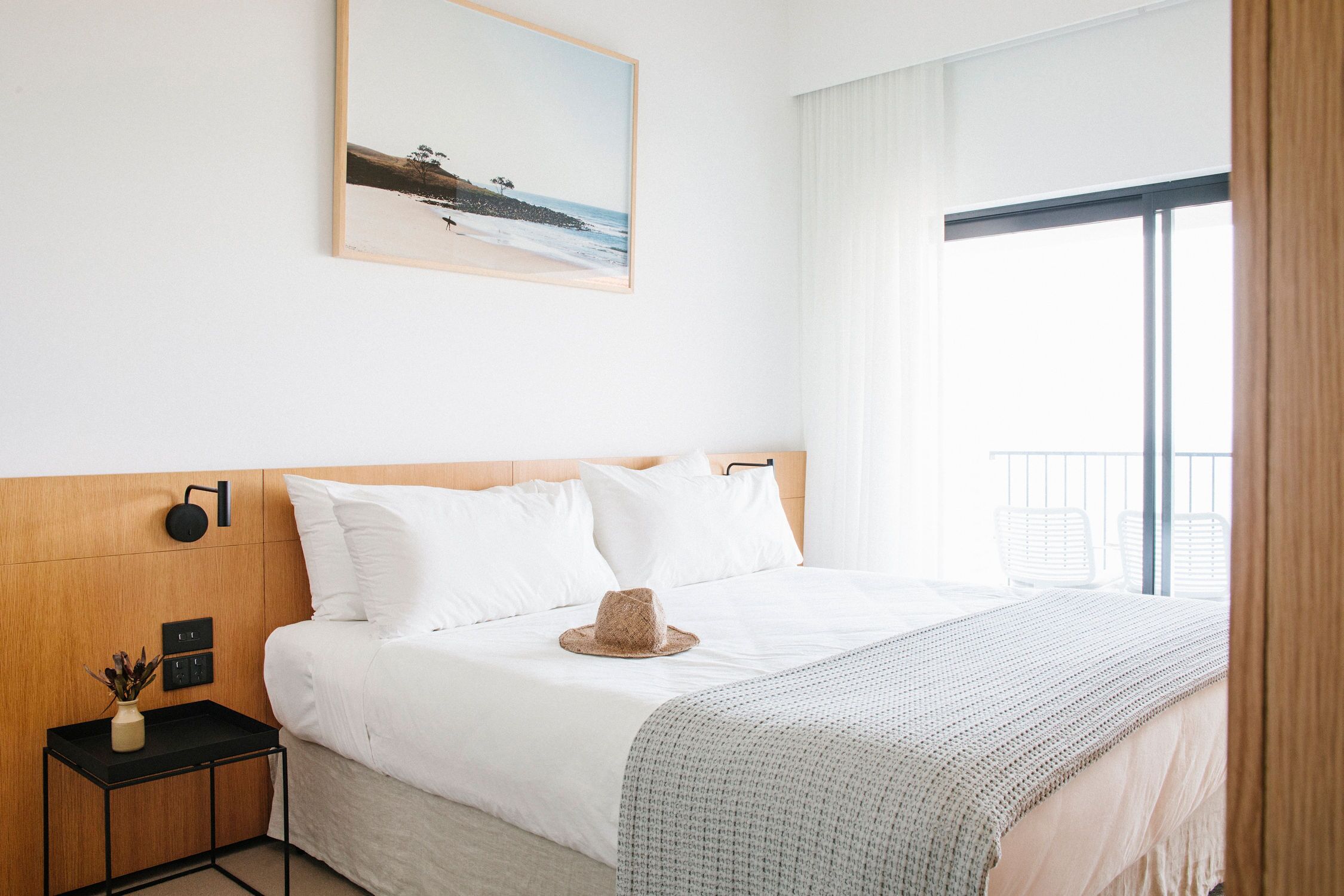 A hotel room with a bed, a nightstand, a wall-mounted lamp, a framed picture, and a window with curtains.