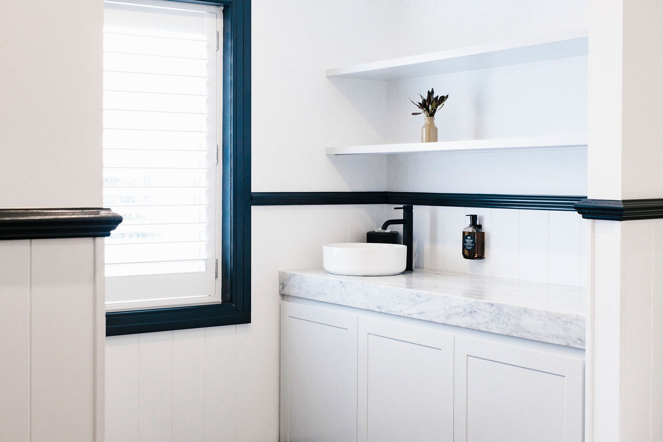 A bathroom with a white sink, a small plant on a shelf, and a window with blinds.
