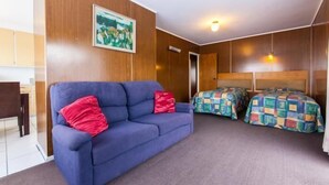 A compact room with a blue sofa, two beds with colorful bedding, a wooden wall, and a painting. - Sunset Lodge Motel (Auckland)