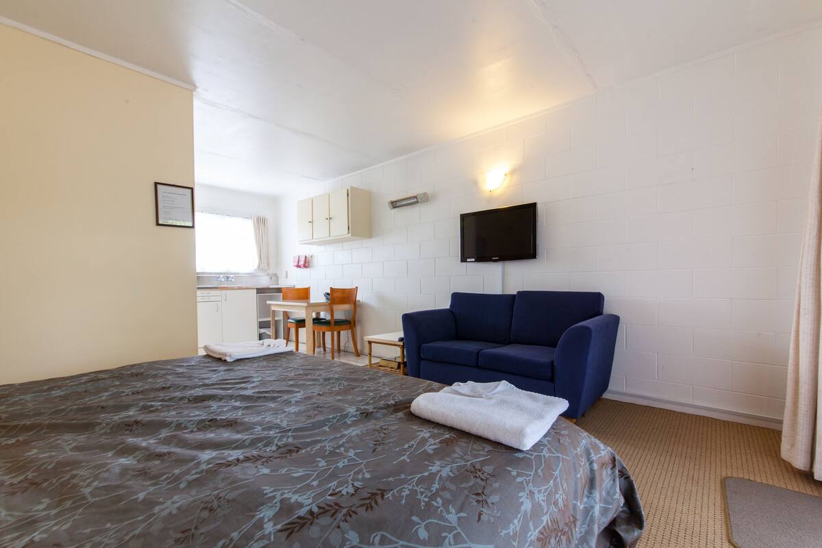 Una habitación de hotel con una cama, un sofá azul, un televisor de pantalla plana en la pared y una kitchenette al fondo.