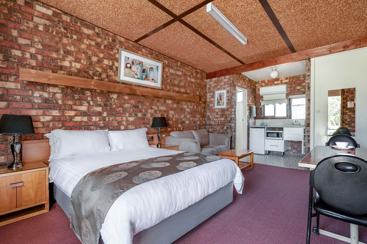 Habitación de hotel con pared de ladrillo, cama con colcha estampada, escritorio con silla y kitchenette.