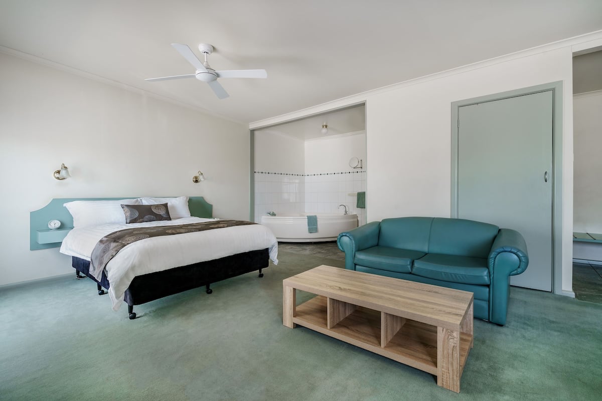 Un dormitorio con cama, una mesa de centro de madera, un sofá color turquesa, un ventilador de techo y un baño con bañera y ducha.